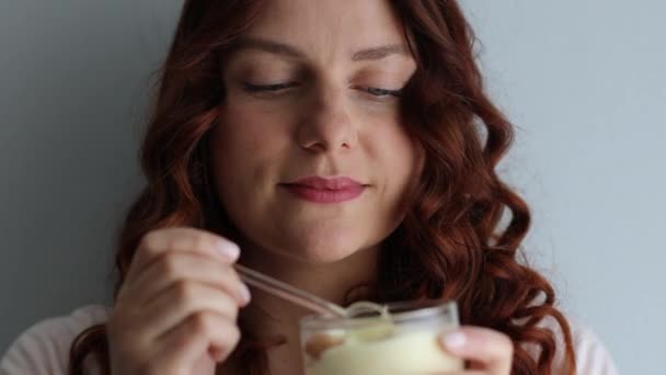 Позитивна кучерява біла дівчина їсть смачний вершковий десерт, насолоджуючись смаком на сірому фоні
 - Кадри, відео