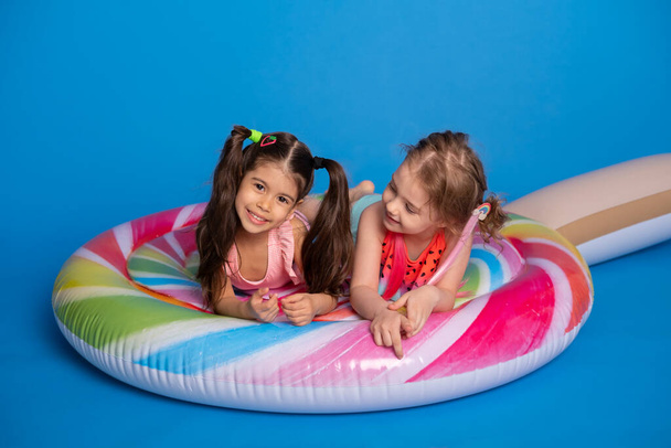 two happy child girl in swimming suit lying on colorful inflatable mattress lollipop on blue background. - Photo, Image