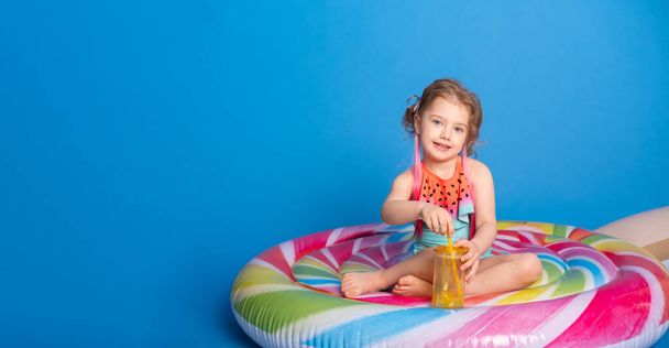 linda niña en traje de baño bebiendo jugo de naranja sentada sobre colorido colchón inflable sobre fondo azul. banner. - Foto, imagen