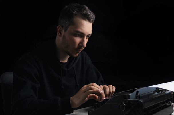 Fiatal jóképű férfi fekete kapucnis gépel egy írógépen, fekete háttérrel, alacsony kulccsal. - Fotó, kép