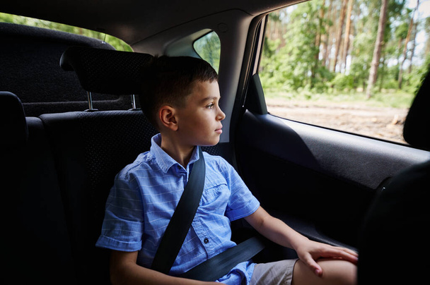 Zapięty nastoletni chłopiec siedzący na fotelu przypominającym samochód bezpieczeństwa i wyglądający przez okno podczas podróży samochodem - Zdjęcie, obraz