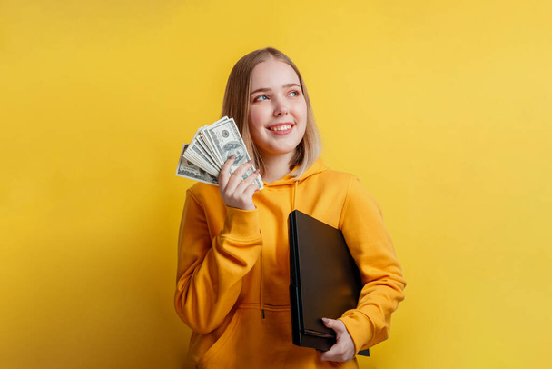 portrait of young exciting smiling pretty woman holding a stack of money dollar bills and laptop. Teenager Girl dreaming of money, earnings online,lottery win isolated on yellow color background - Foto, Imagen