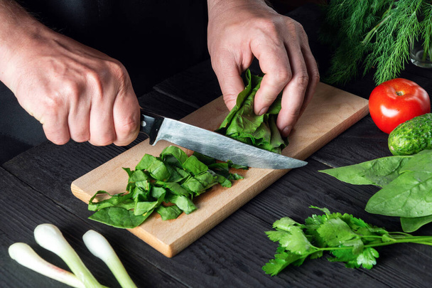 Un chef prepara una ensalada de espinacas en la cocina de un restaurante. Primer plano de las manos de un cocinero cortando verduras con un cuchillo en una tabla de cortar. - Foto, Imagen