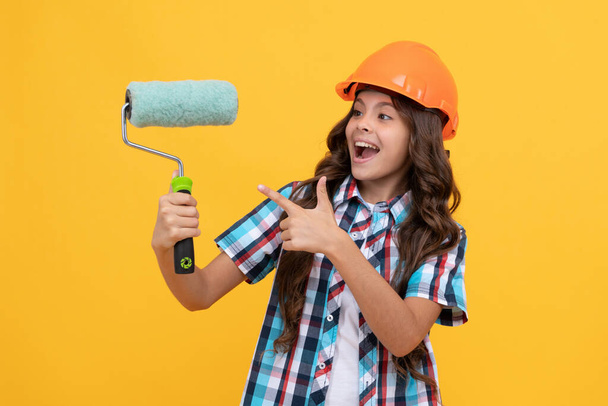 szczęśliwy nastolatek dziewczyna z kręconymi włosami w kasku budowlanym wskazując palcem na wałku farby, remont - Zdjęcie, obraz