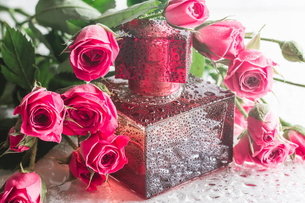 Бургундские квадратные парфюмерные бутылки на глянцевой поверхности с тонкими розовыми розами и каплями воды. Eau de Feltte. - Фото, изображение