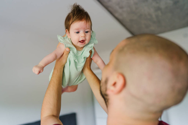 Homme caucasien tenant sa petite fille enfant fille haut dans l'air à la maison dans la chambre dans la journée d'été - concept de famille de la fête des pères de la parentalité de vraies personnes - Photo, image