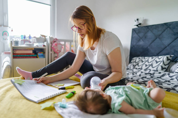 新生児の赤ちゃんの娘をケアしながら、家庭学習でベッドの上に座って千年の白人女性の母親のフロントビュー-教育親の概念現実の人々 - 写真・画像