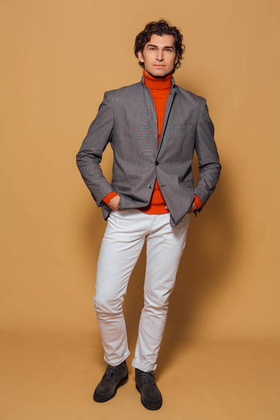 Fashion portrait of a tall handsome man dressed in orange turtleneck, white jeans and grey jacket posing on the beige background - Fotoğraf, Görsel