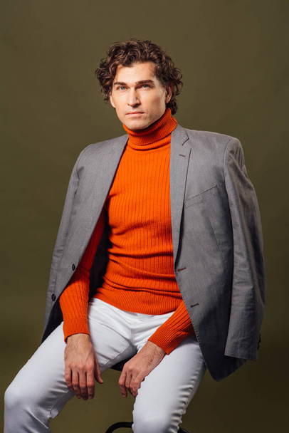 Portrait of a tall handsome man dressed in orange turtleneck, grey jacket and white jeans posing on the green background - Foto, Imagen