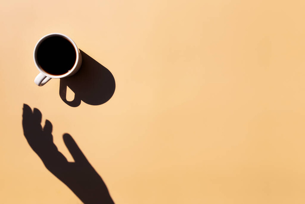 Taza blanca y platillo de café expreso negro desde la vista superior con sombra de sol y mano sosteniendo o tocando la taza sobre fondo de terracota beige amarillo. Foto de alta calidad - Foto, imagen