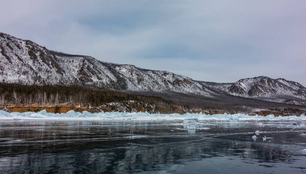Des éclats de glace sont éparpillés sur la surface lisse et brillante du lac gelé. Blocs de hummocks près du rivage. Chaîne de montagnes enneigée contre le ciel bleu. Réflexion. Baïkal - Photo, image