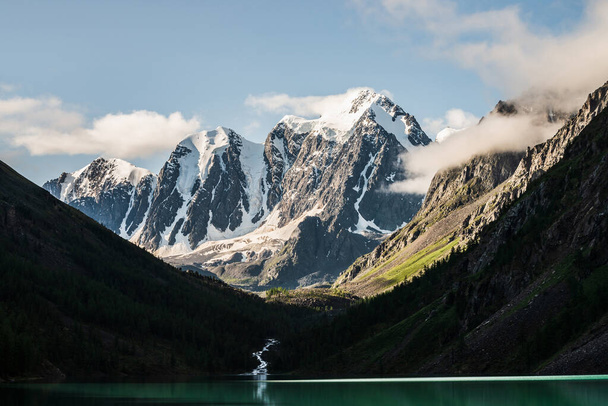 Piękny duży lodowiec, skaliste góry śnieżne, las iglasty na wzgórzach, górskie jezioro i potok górski pod błękitnym niebem z chmurami. Atmosferyczna sceneria alpejska z niskimi chmurami na wysokich stromych zboczach. - Zdjęcie, obraz