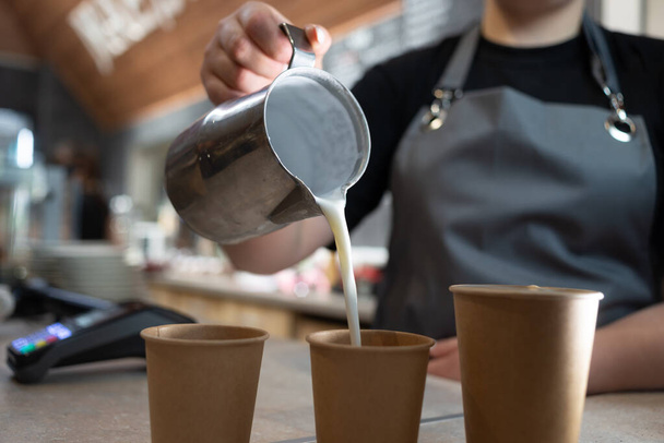 Egy barista nő közelről tejet önt a pohárba egy fémkancsó kávéjával. Három papírpohár forró itallal. A cappuccino készítésének folyamata. - Fotó, kép