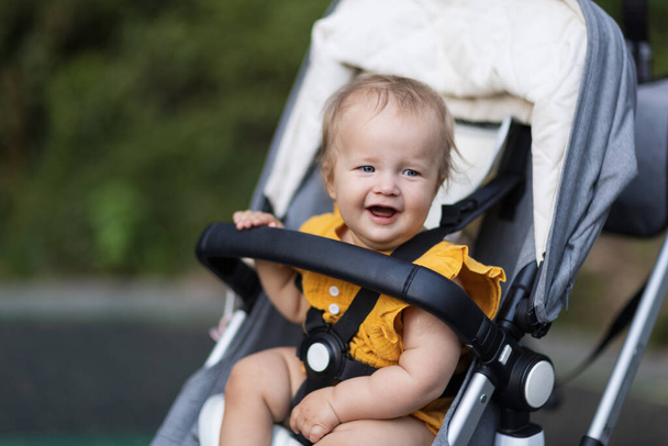 Sweet caucasian baby girl ten months old sitting in stroller outdoors. Little child in pram. Infant kid sits in pushchair. Summer walks with kids. Family leisure with little child. - Фото, зображення
