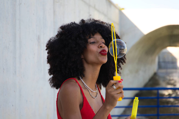 krásná afro americká žena dělá mýdlové bubliny v parku. Žena se skvěle baví s bublinkami, a proto se usmívá do kamery. Pojem štěstí - Fotografie, Obrázek