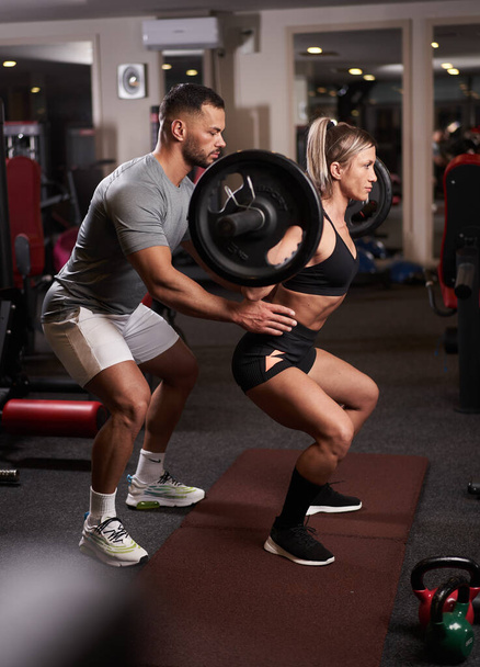 Athletic woman doing barbell squats assisted by her fitness trainer - Foto, Bild