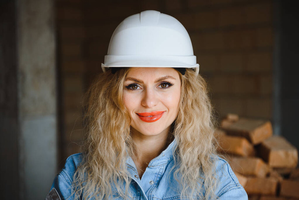 Attractive female construction worker in hardhat. Confident young specialist in checkered blue shirt in jeans standing in empty room. Interior design and renovation service. - Foto, Bild
