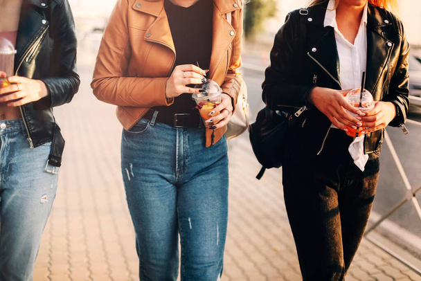 Happy young women with takeaway drinks walking on pavement and smiling on sunny weekend day on city street - Foto, Imagen