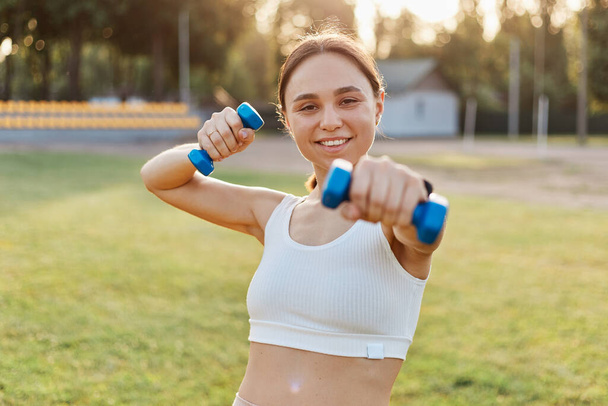 Portrait of happy satisfied smiling female wearing white top holding out dumbbell to camera, training arms, doing exercises for biceps and triceps, expressing positive emotions while doing sport. - Foto, Imagem