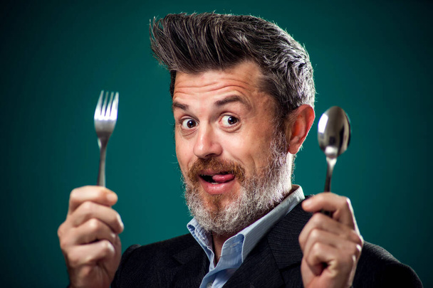 Ένα πορτραίτο ενός πεινασμένου γενειοφόρου άντρα με κοστούμι που κρατάει κουτάλι και πιρούνι.  - Φωτογραφία, εικόνα