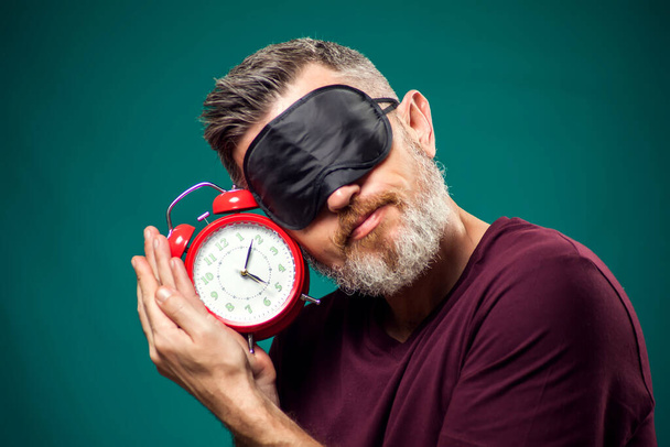 Surprised man in red t-shirt and sleep mask on head holding red alarm clock. Lifestyle and bed time concept - Foto, Bild