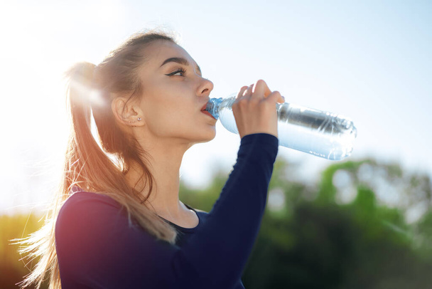 Portrait of young beautiful woman wearing blue sportswear drinking water at park - Photo, Image