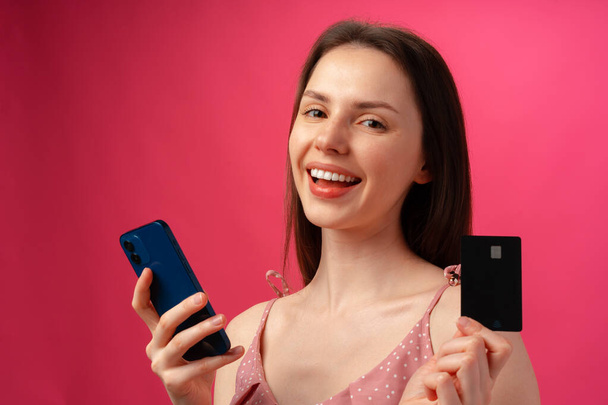 Smiling young woman holding smartphone and credit card against pink backgorund - Foto, afbeelding