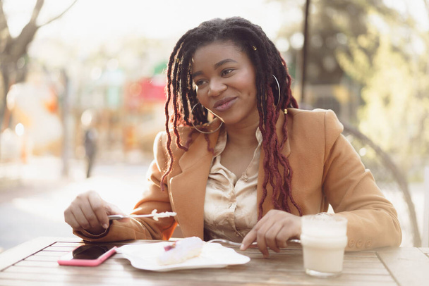 Smiling african american woman drinking coffee and eating dessert in outdoor cafe - Fotoğraf, Görsel