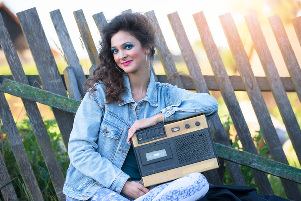Uma linda garota do campo em roupas brilhantes se senta em um banco de madeira com um gravador de cassetes. Mulher no estilo dos anos 90. - Foto, Imagem
