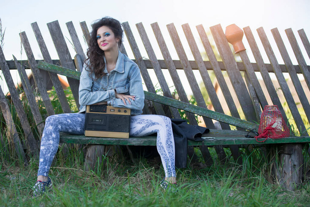 Μια όμορφη χωριατοπούλα με φωτεινά ρούχα κάθεται σε ένα ξύλινο παγκάκι με ένα κασετόφωνο. Γυναίκα στο στυλ της δεκαετίας του '90. - Φωτογραφία, εικόνα