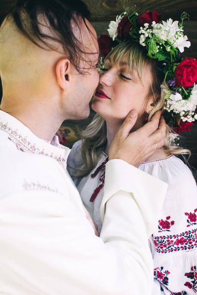 Young people are posing in traditional folk costumes. Cossacks man and woman in wreaths and embroidered shirts. Reconstruction of vintage traditions and customs. Guy and girl kiss - Photo, Image