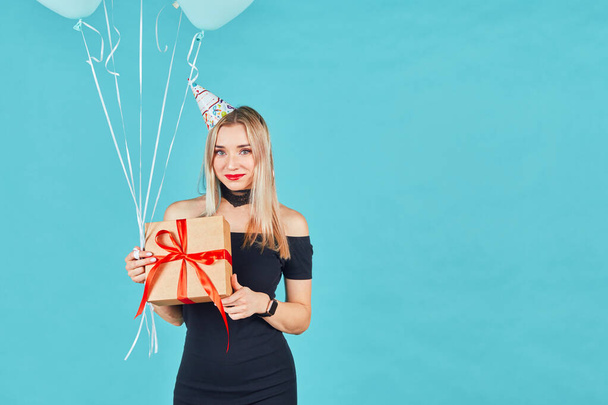 Beautiful smiling woman in black dress and birthday hat holding gift box and pastel air balloons against blue background. Happy young girl celebrating birthday party. - Photo, image
