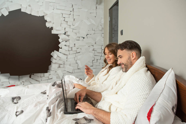 Handsome couple in bathrobe lying on the bed using notebook - Φωτογραφία, εικόνα