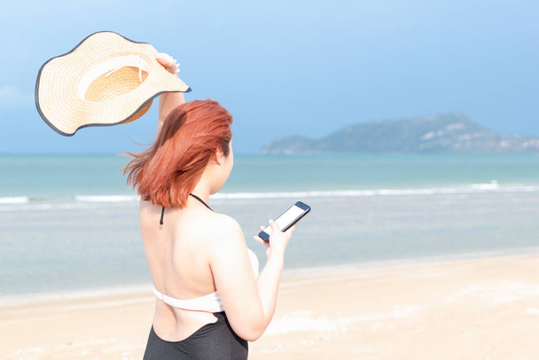 Frau im Badeanzug, linke Hand mit Hut, rechte Hand mit Handy am Strand. Leere Handy für Attrappe. Selektiver Fokus am Telefon - Foto, Bild