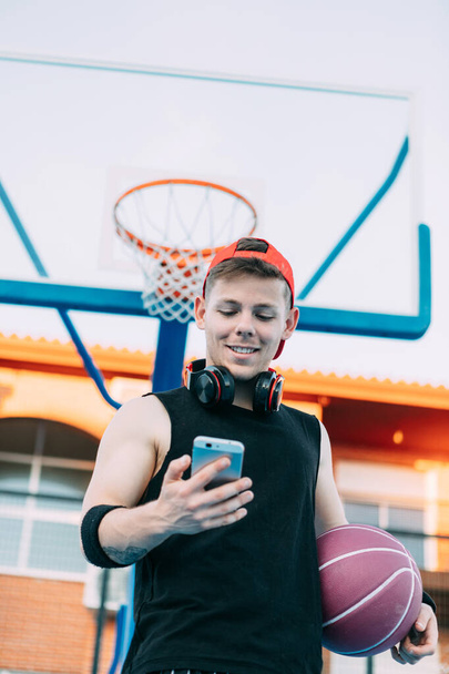 Aantrekkelijke man basketbalspeler met een bal kijkt naar zijn mobiele telefoon en glimlacht tijdens het nemen van een pauze - Foto, afbeelding