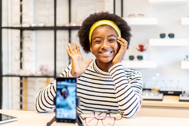 若い女性アフリカ系アメリカ人起業家は、携帯電話にファッションvloggerショーグラスであり、眼鏡店でオンラインライブストリーミングそれを販売笑顔 - 写真・画像