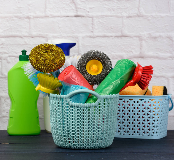 esponjas, cepillos de plástico y botellas de detergentes sobre una mesa de madera azul. Artículos de limpieza del hogar sobre fondo de pared de ladrillo blanco - Foto, imagen