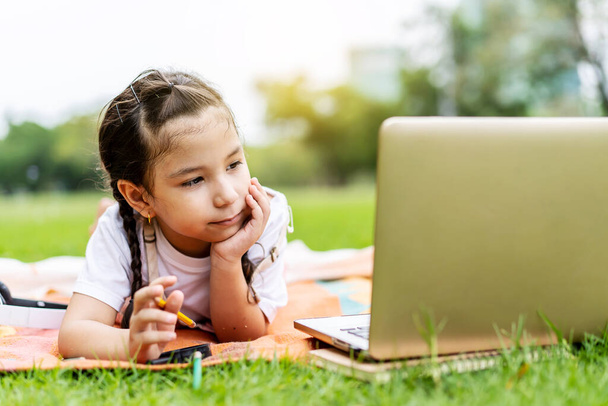 Szczęśliwe dziecko dziewczynka uśmiecha się, rozmawia, uczy się i dobrze się bawi patrząc na laptopa w parku letnim. Koncepcja kształcenia on-line - Zdjęcie, obraz