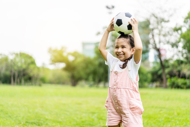 Smiling little girl standing holding the soccer ball at green football field in summer day. Portrait of little girl athlete playing with a ball at stadium. Active childhood concept. Copy space - Fotografie, Obrázek
