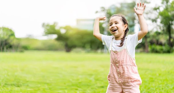 Ευτυχισμένο όμορφο κοριτσάκι χαμογελά και σηκώνει το χέρι της, ενώ στέκεται στο πάρκο, Θετική θηλυκό παιδί απολαμβάνει ζεστή μέρα στο πάρκο φθινόπωρο - Φωτογραφία, εικόνα