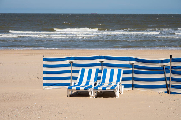 Vacías tumbonas azul-blancas en la playa de arena amarilla en la pequeña ciudad belga De Haan o Le Coq sur mer, destino de vacaciones de lujo, vacaciones de verano - Foto, imagen