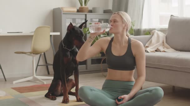 Steadicam of fit Valkoihoinen nainen yllään urheiluvaatteet, istuu jalat ristissä lattialla kotona, juo vettä pullosta, aikuinen dobermanni koira istuu lähellä - Materiaali, video