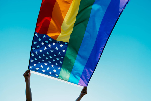 Nahaufnahme einer Person, die eine regenbogenfarbene US-Flagge am blauen Himmel schwenkt, mit der Sonne im Hintergrund - Foto, Bild