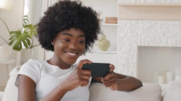 Happy relaxed millennial mixed race girl holding smartphone looking at cellphone screen laughing enjoying using mobile apps for shopping having fun playing games chatting in social media sit on couch - Felvétel, videó