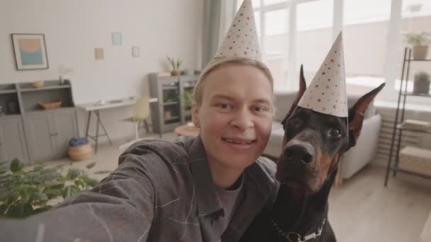 Handheld POV młodej białej kobiety i czarnego psa Dobermana w kapeluszach party, robienie selfie w domu, właściciel uśmiechający się i całujący zwierzaka - Materiał filmowy, wideo
