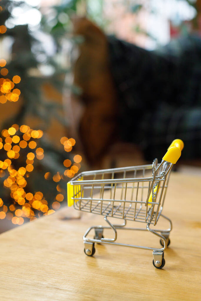 New Year and Christmas background. Small grocery cart stands on wooden table against background of bokeh garlands on christmas tree and sofa - Φωτογραφία, εικόνα
