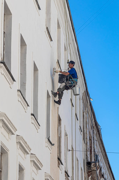 Μόσχα, Ρωσία, 06.17.2021. Ένας βιομηχανικός ορειβάτης κρέμεται στα σχοινιά ασφαλείας σε ύψος και σφραγίζει παρατυπίες στον τοίχο με μια σπάτουλα. - Φωτογραφία, εικόνα