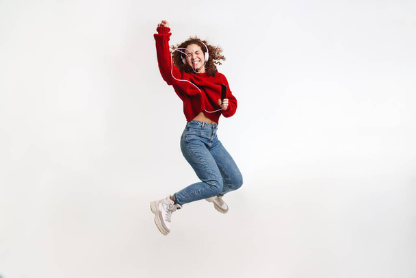 Joyful girl jumping while listening music with cellphone and headphones isolated over white background - Foto, Imagem