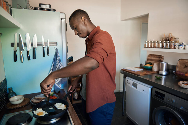 Young African man cooking eggs in a frypan on his kitchen stove while making breakfast in the morning - Fotoğraf, Görsel