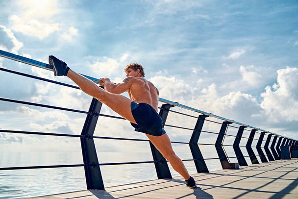 Young muscular tattooed man doing exercises, stretching early in the morning on pier by the sea - Foto, imagen